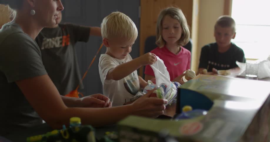 Several children and an adult gathered at a table watching another boy open a present
