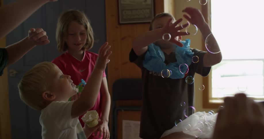 A girl blows bubbles with a bubble blower as other children swipe at the bubbles and an adult takes pictures