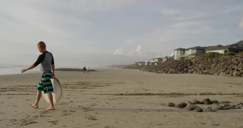 A boy in striped shorts walks toward the water on a beach