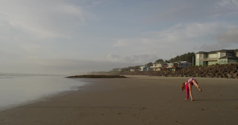 A little girl performing a cartwheel on a beach under cloudy skies