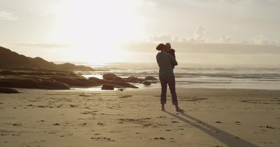 A woman and baby stand on a beach at sunset