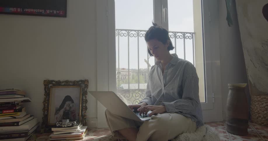 Woman sits on pad on floor typing on laptop propped in lap