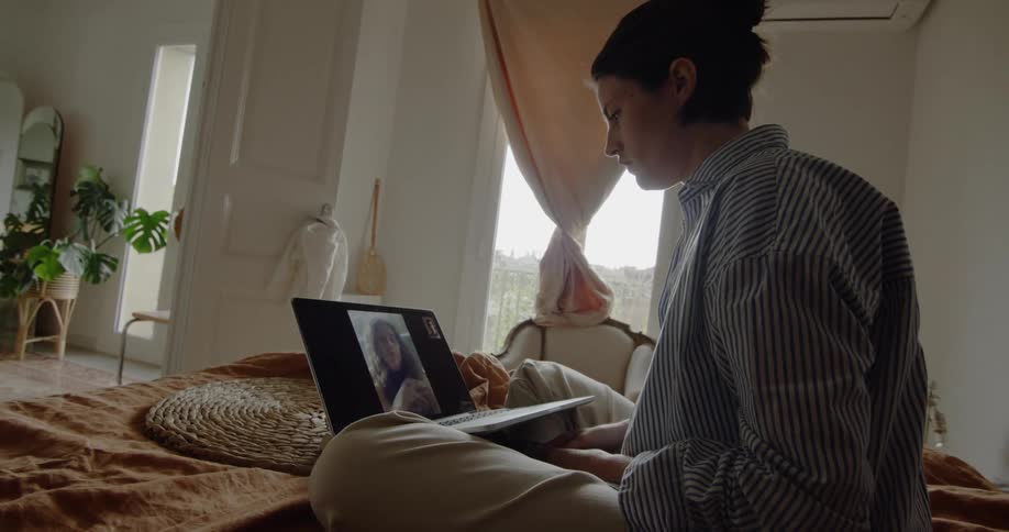 A woman sits on her bed and watches a video on her laptop