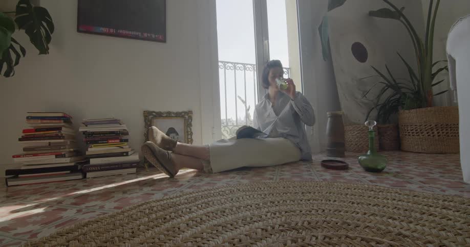 A woman sits on the floor and flips through a book and then takes a drink of water