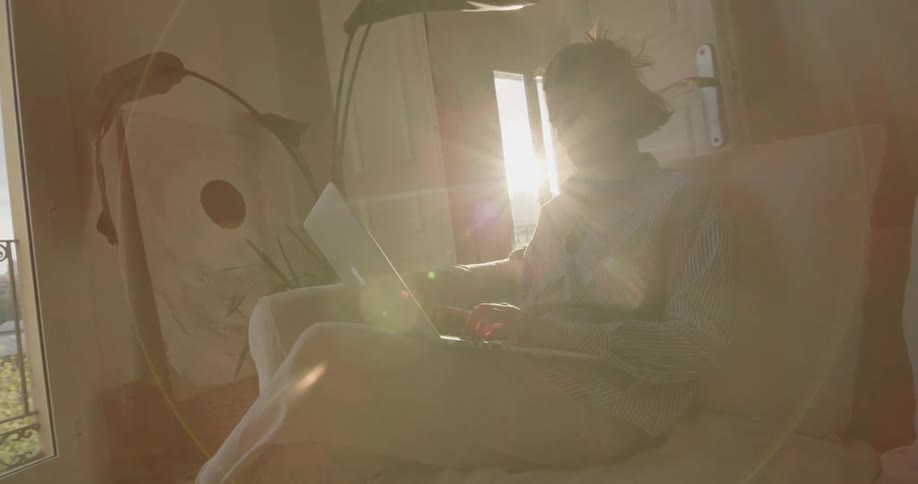 Sunlight shines behind a woman using a laptop on a living room sofa