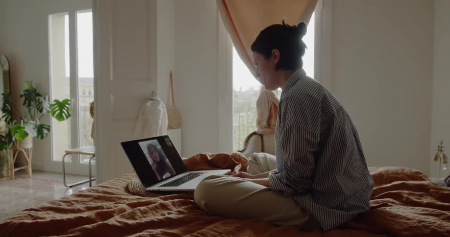 A woman sits on her bed and video chats with another woman on a laptop