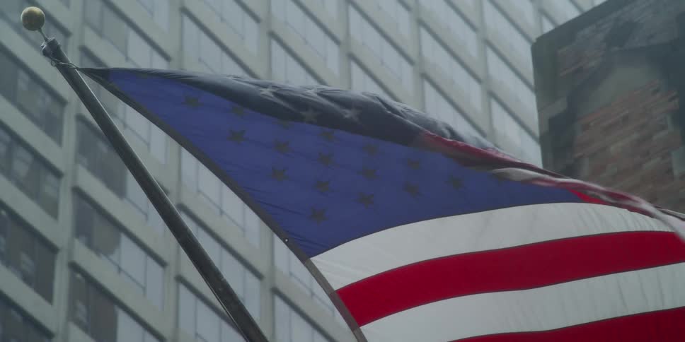 An american flag blows in the wind with city buildings and windows behind it