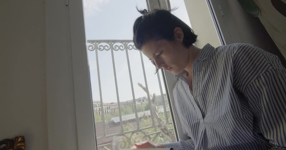 Female sits next to a window reading a book while holding a pencil in her left hand