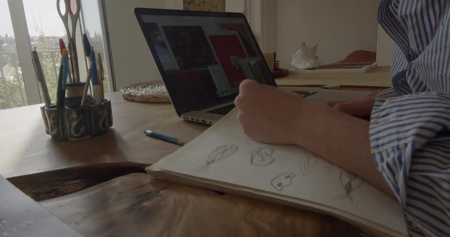 A person sits in front of a laptop sketching with a pencil in a drawing pad