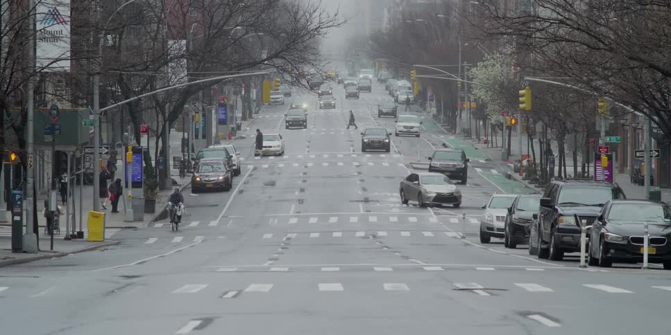 Cars turn onto and travel on a city street with people walking and riding bicycles on a foggy day