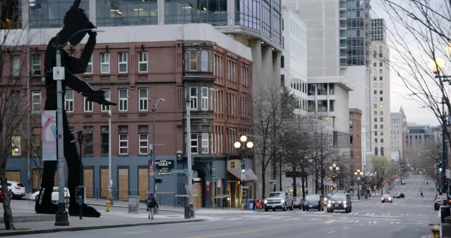 Cars travel on a city street and people walk on the sidewalk where a large art sculpture of a hammering blacksmith stands
