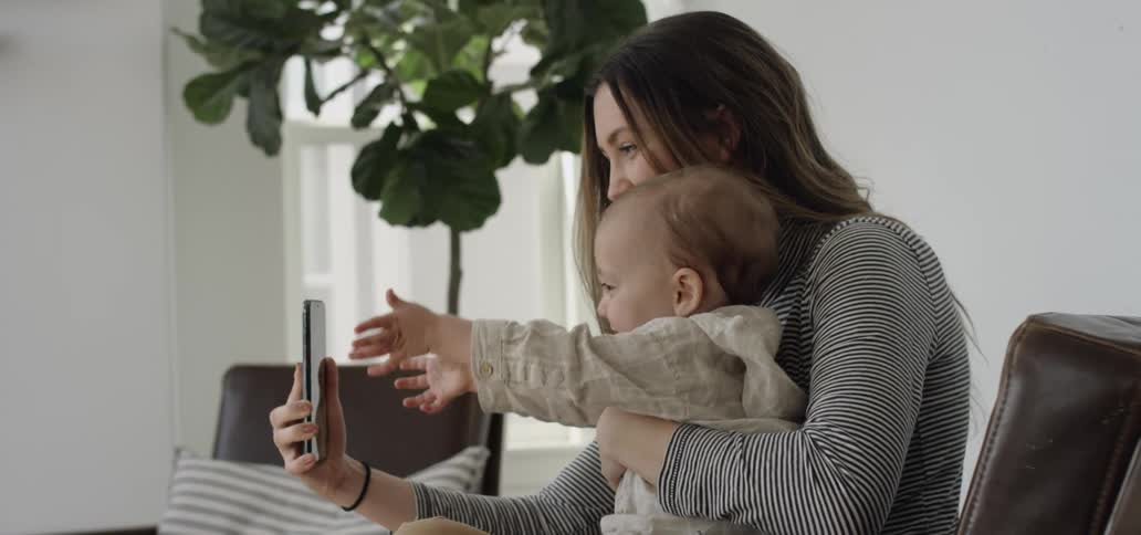 A woman holds a toddler and uses a cell phone for a video call