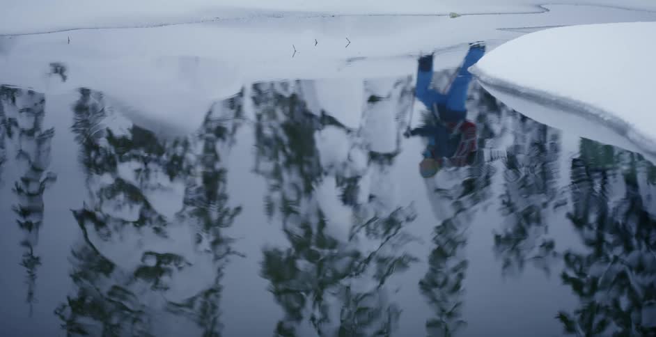 Skier moves past rippling reflective water bordered by snowy shore