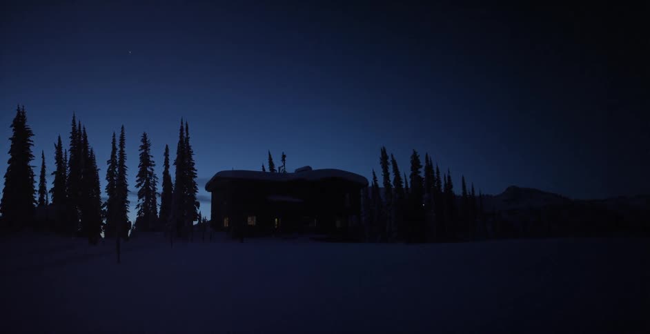 A man walking across a snowy field in front of a cabin in the darkness of early morning