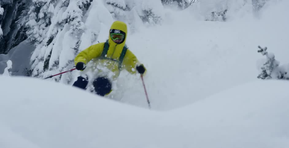 A skier in a yellow coat speeds down a snowy mountainside