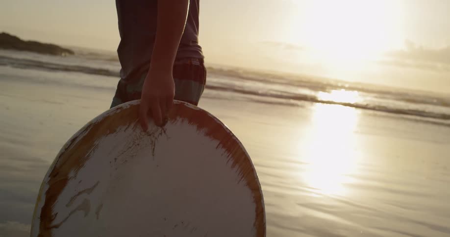 A boy walks down a beach while carrying a disk ski at sunset