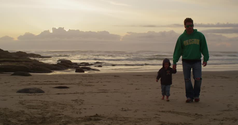 A man holding a little boy's hand as they walk away from the shoreline on a chilly, cloudy day