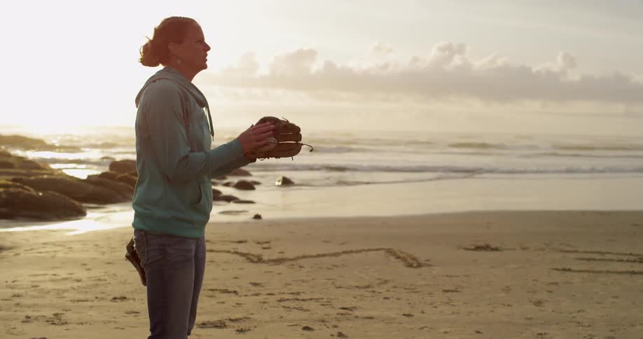 A woman wearing a baseball mitt throwing a baseball to someone and catching it as it gets thrown back to her on the beach under cloudy skies