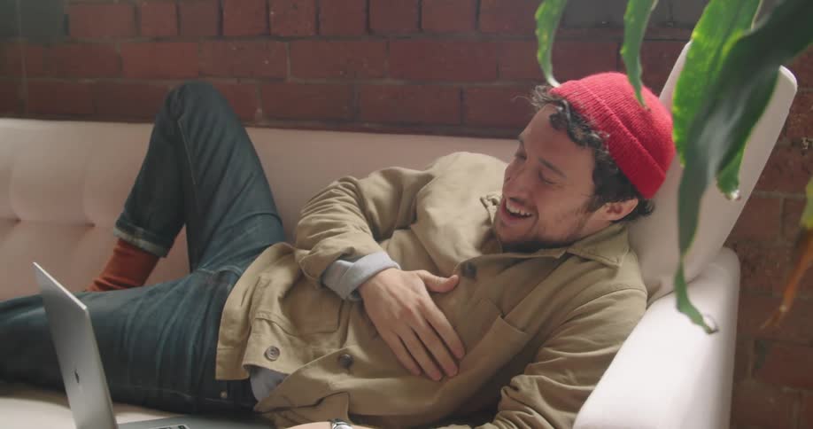 A man lays on the couch and laughs while talking to someone using his laptop