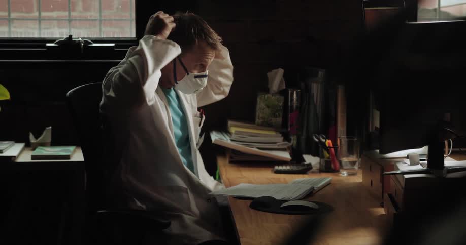 A doctor sitting at a desk puts on a face masks and gets up to leave