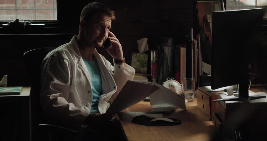 A doctor sits at a desk and goes through notes while talking on a phone