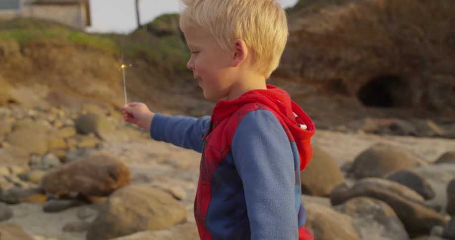 A small boy walking over a sandy area with a sparkler near boulders and cliffs