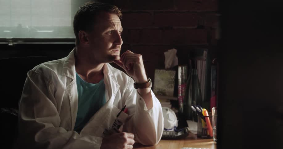 A male doctor is sitting at his desk with a concerned look on his face and rests his chin on his hand while thinking