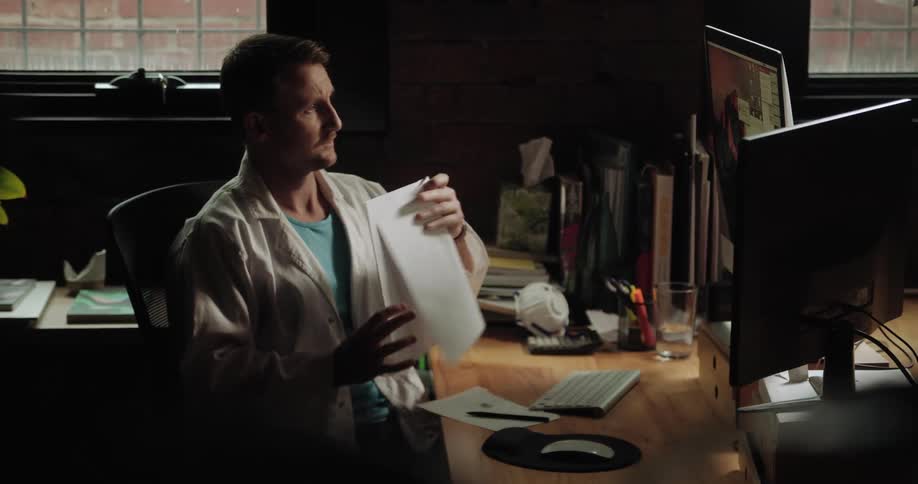 A man in a white lab coat sits at a desk reading pages then typing on a keyboard