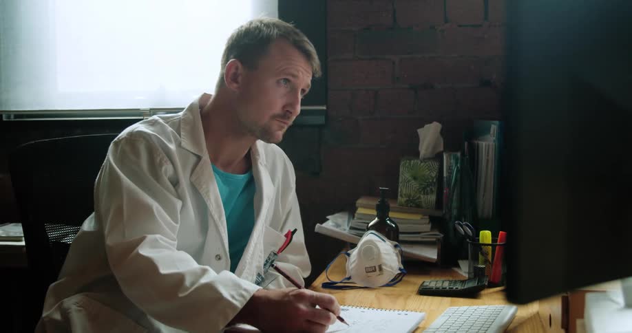 Doctor sitting at the desk looks at the computer display and takes notes in the office