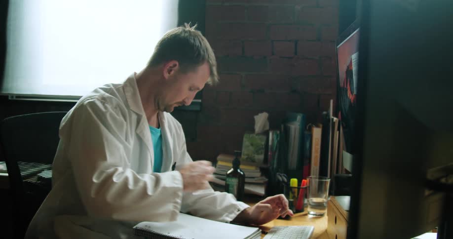 A doctor looks at a computer monitor and types on a keyboard then gets a pen to write in a notebook
