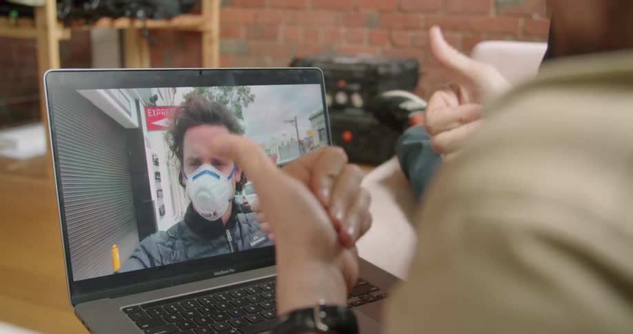 Man sitting on the sofa makes a video call with his friend wearing a face mask on the laptop