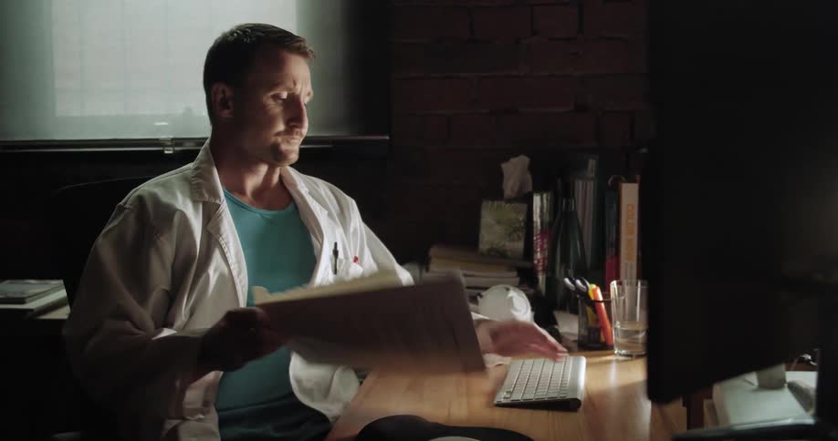 A man sits at his desk and looks at his computer and paperwork