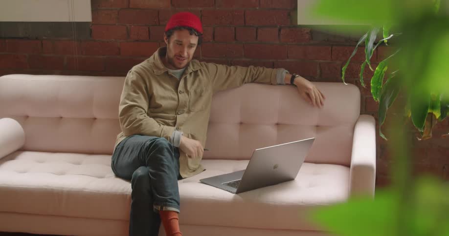 A man talks while sitting on a couch and looking at a laptop