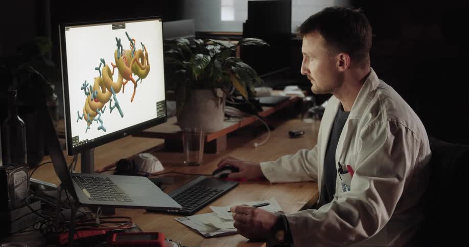 A man takes notes at his desk while viewing a graphic on his computer monitor