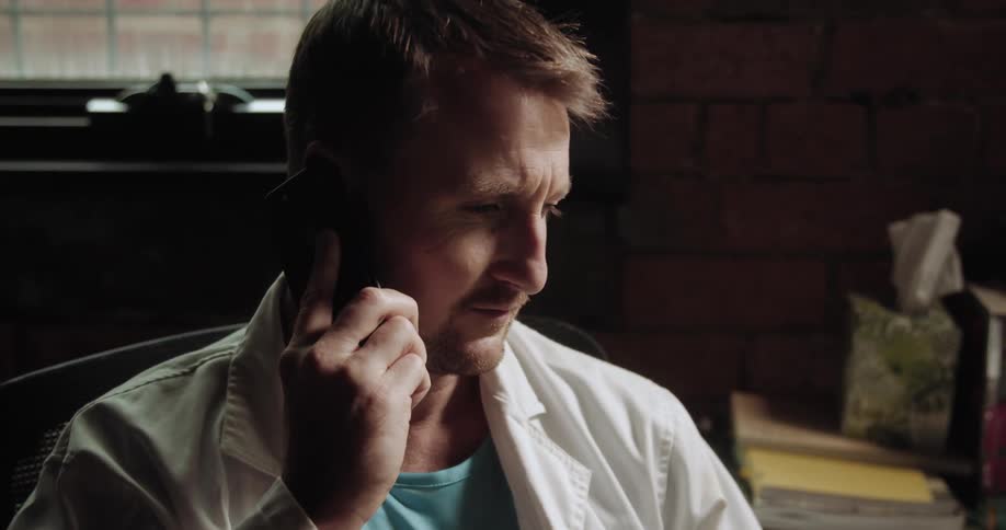 A doctor in a lab coat sits at a desk while talking on a smartphone