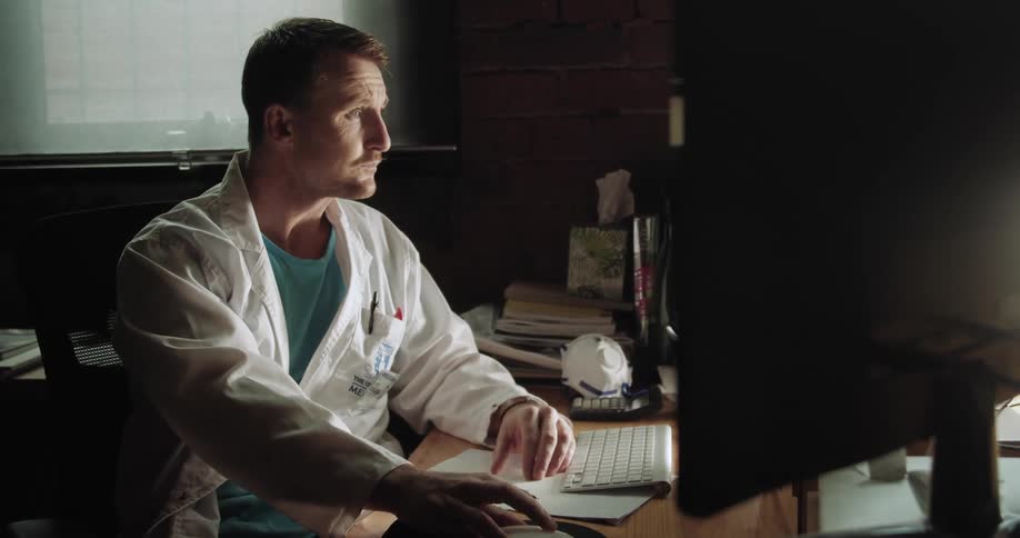A male doctor sits at a desk in front of a computer and writes something down