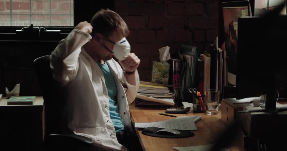 A doctor putting on a face mask while sitting at his desk before rising to leave the room