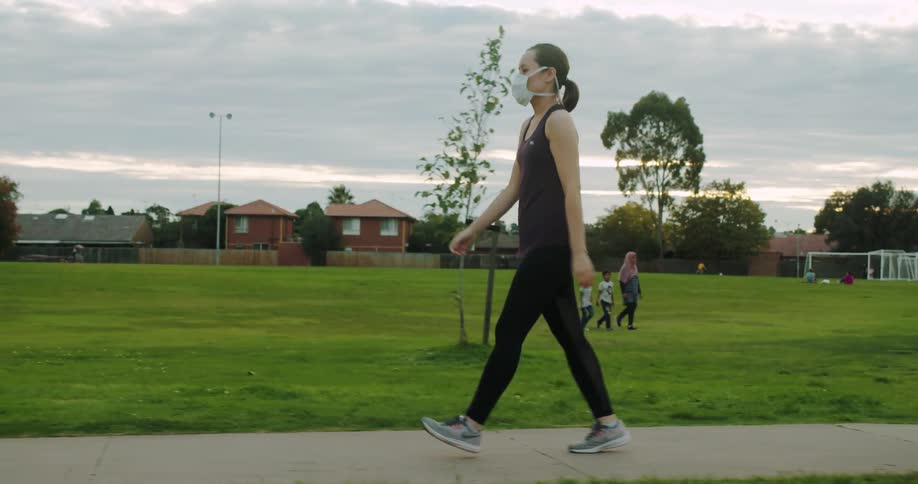 A woman wears a medical mask walking down a suburban sidewalk