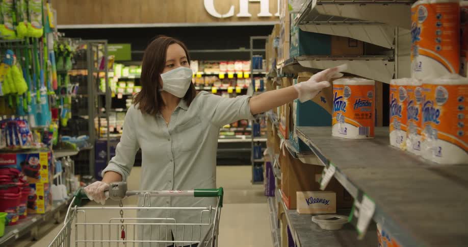 A woman wearing a mask pushes her grocery cart down the aisle and picks up paper towels from the shelf