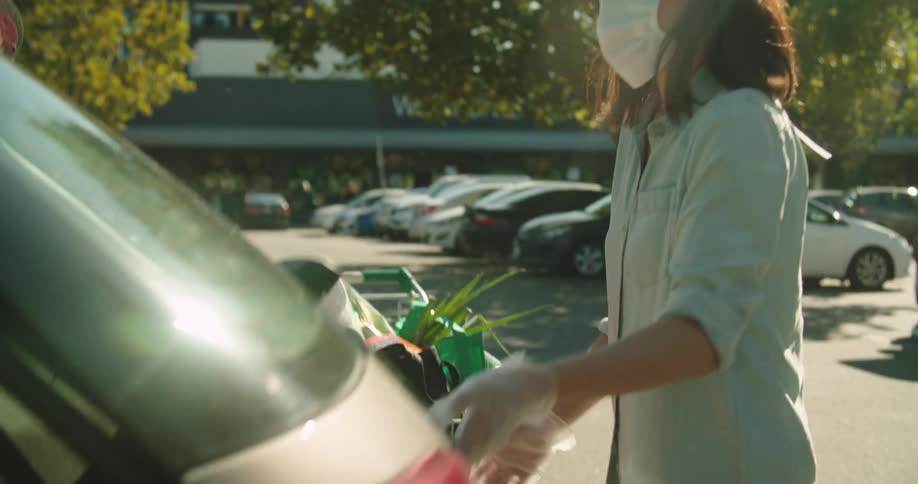 Woman wearing gloves and face mask loads groceries into her vehicle