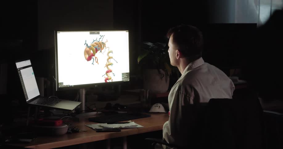 A man sits at his desk and writes on paper as he looks at his laptop and desktop computer