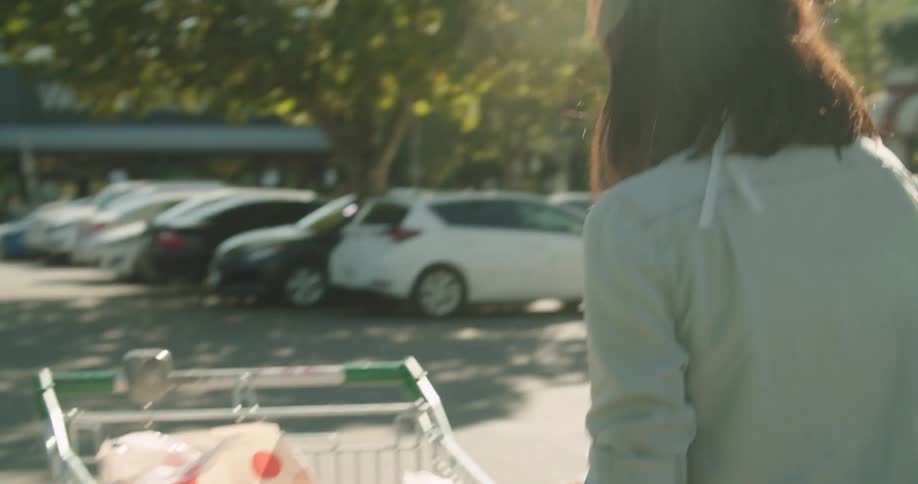 A woman wearing a mask takes bags of groceries out of a cart and puts them in her car