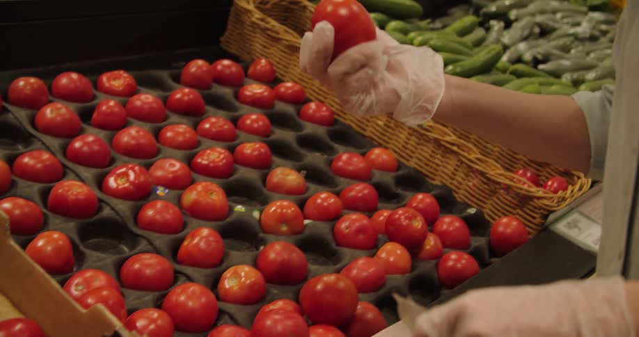 Gloved hands picking up a tomato at produce section in a store