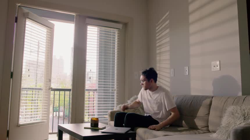 A man sits on a couch using his laptop and then gets up and goes out on his balcony