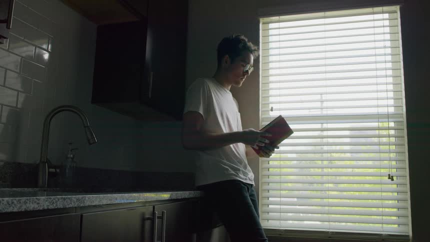 Man reclines on kitchen countertop near window and opens a book