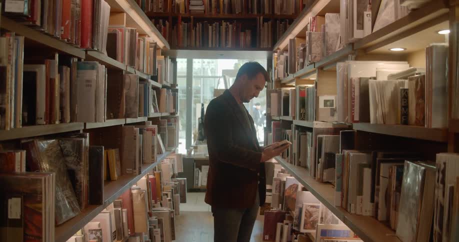 A man walks down an aisle of books and takes one of the shelf to look at it