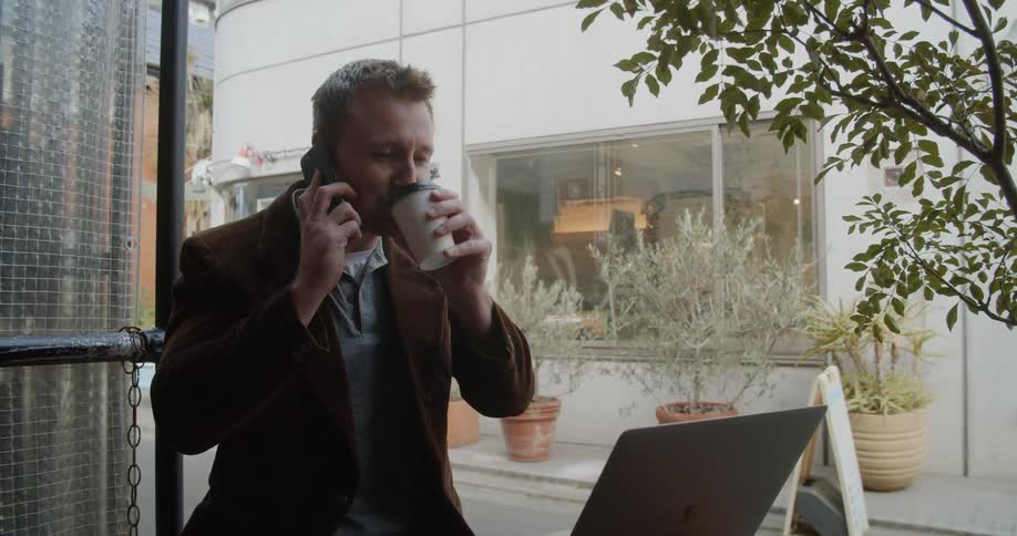Man sits outdoors talking on phone while studying laptop and drinking coffee