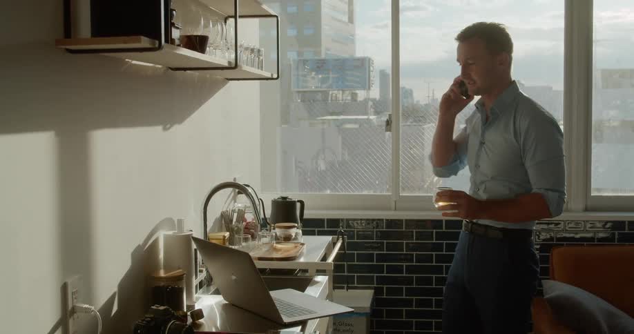 A man standing near the sink in an apartment while talking on the phone and drinking