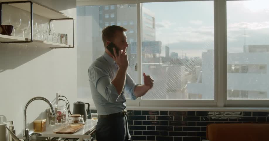 Man stands in kitchen of apartment overlooking city talking on cell phone