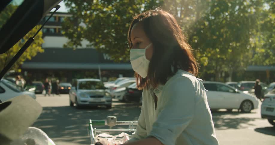 A woman wearing a surgical mask loads bags into her car from a shopping cart